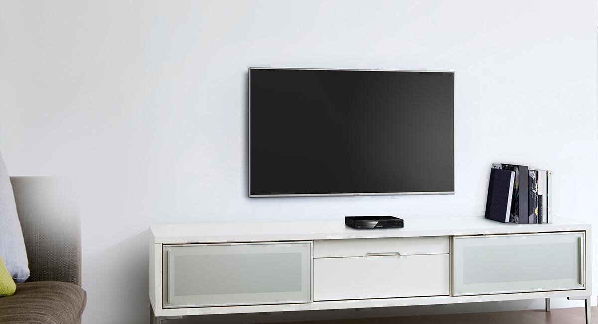 Na białej szafce znajduje się czarny odtwarzacz Blu-ray, a nad nim wisi płaski telewizor. Obok odtwarzacza leżą książki lub czasopisma.