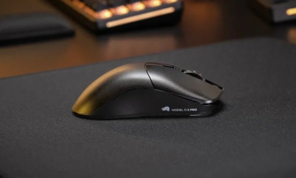 Czarna mysz komputerowa z napisem \'MODEL O 1 PRO\' na boku, umieszczona na ciemnej podkładce. W tle widoczna jest klawiatura.