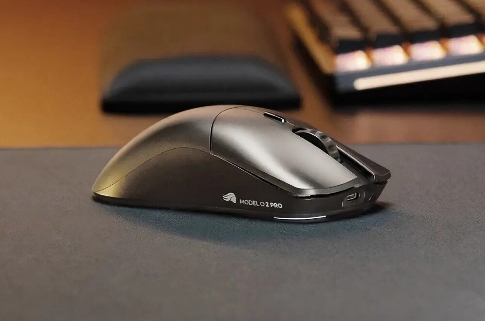 Czarna mysz komputerowa z napisem \'MODEL O 2 PRO\' na boku, umieszczona na ciemnej powierzchni. W tle widoczna jest klawiatura i inne elementy biurka.