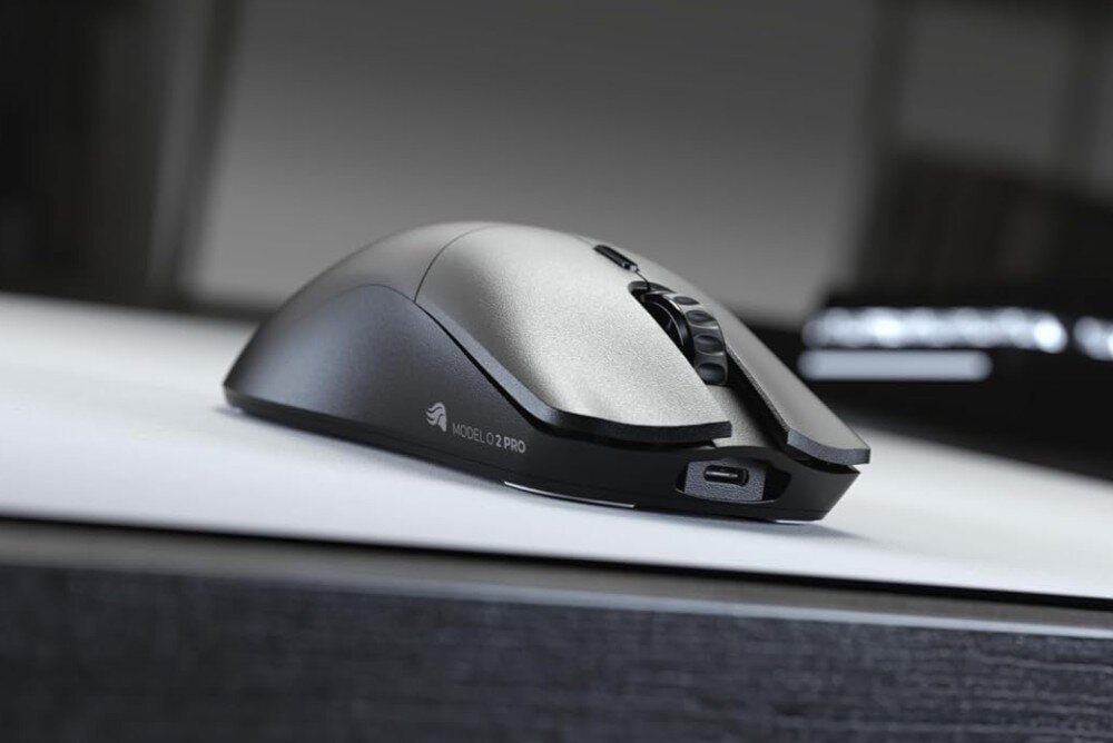 Czarna mysz komputerowa z napisem \'MODEL O 2 PRO\' i logo producenta na boku, leży na białej powierzchni. W tle widoczny jest rozmyty ekran.