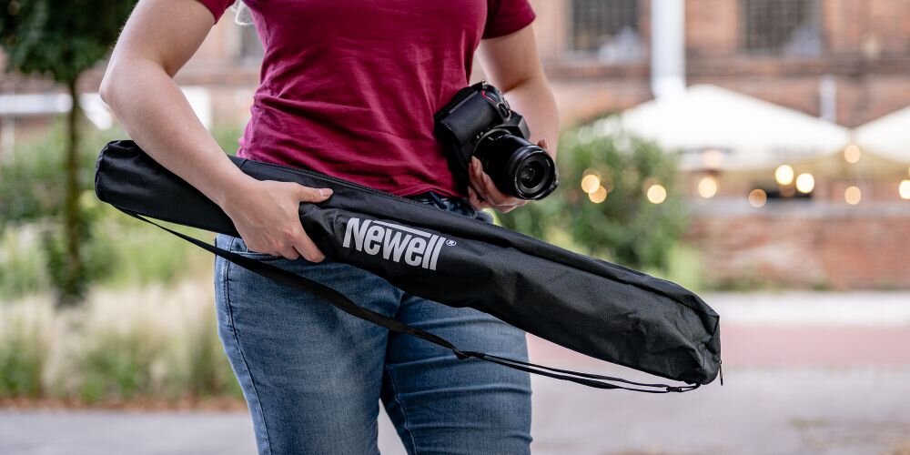 Osoba trzyma czarną torbę z napisem \'Newell\' oraz aparat fotograficzny. W tle widoczne są rozmyte elementy miejskiego krajobrazu.