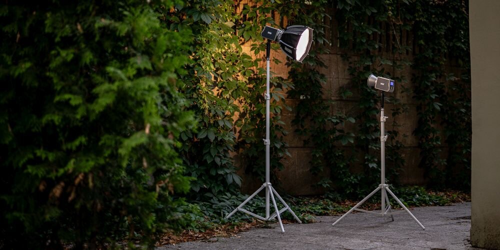 Na obrazie znajdują się dwa statywy z zamontowanymi lampami, ustawione na tle zielonej roślinności. Jeden z nich oświetla ścianę, tworząc ciepłe światło.
