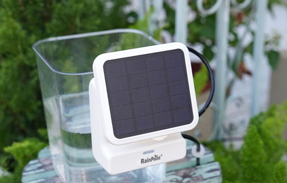 Zestaw do nawadniania RAINPOINT HTP115FR + HWG023WBRF, 10 roślin, mikronawadnianie, wilgotność, system, Biały panel solarnymstoi na zewnątrz obok szklanego naczynia z wodą.