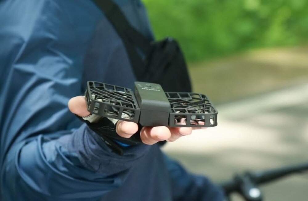 Dron HOVERAIR X1 Combo Plus Dłoń trzymająca złożony czarny dron na tle kierownicy roweru