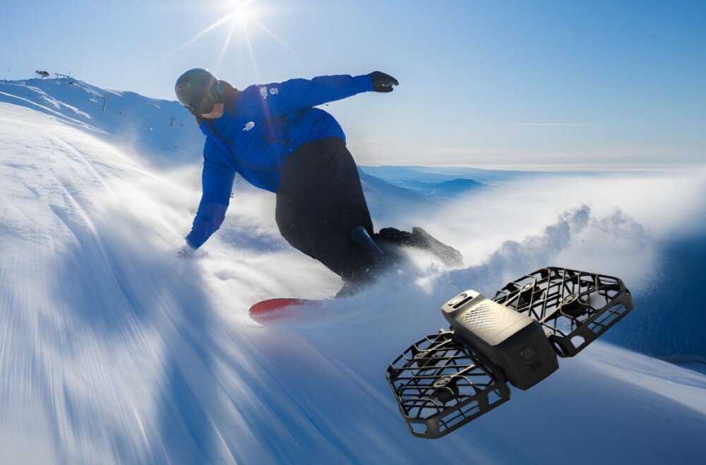 Dron HOVERAIR X1 Promax Snowboardzista w niebieskiej kurtce zjeżdżający po ośnieżonym stoku w pełnym słońcu, śledzony z boku przez niewielki dron w ochronnej kratownicowej obudowie, opis
