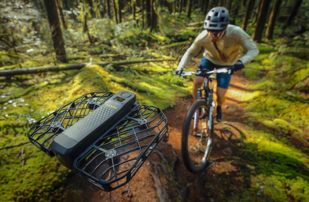 Dron HOVERAIR X1 Promax Rowerzysta górski jadący w lesie po ścieżce między drzewami, przed nim w powietrzu zawisa prostokątny dron w kratownicowej klatce z widocznym napisem 'HOVER', tryby AI, sterowanie