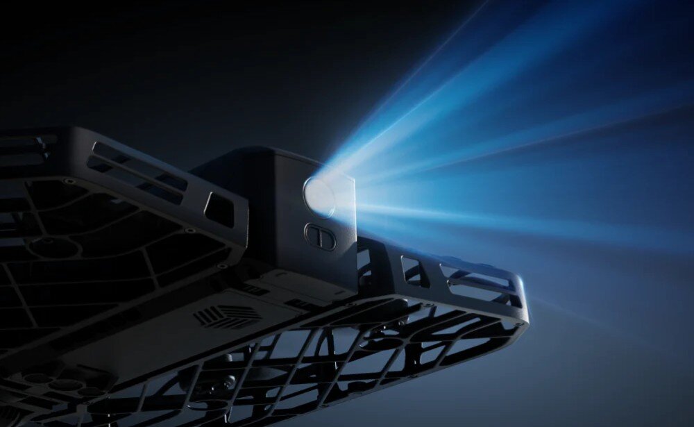 Dron HOVERAIR X1 Promax Ujęcie od spodu na róg prostokątnego drona, z którego przedni reflektor świeci silnym snopem niebieskawego światła na ciemnym tle, konstrukcja, bezpieczeństwo
