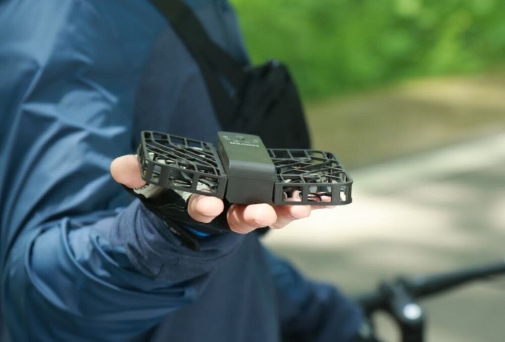 Dron HOVERAIR X1 Promax Dwa ujęcia kolarzy szosowych: po lewej rozmazany z przodu rowerzysta w pomarańczowej koszulce z małym dronem lecącym nad drogą, po prawej inny kolarz na szutrowej drodze, obok którego w powietrzu unosi się podobny dron, bateria, czas lotu, zasięg