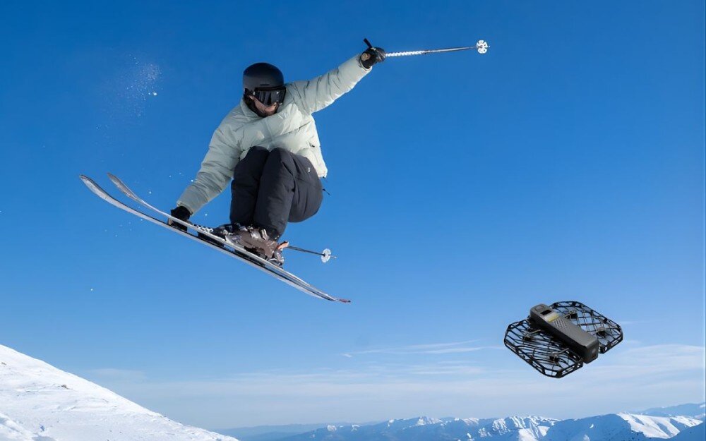 Dron HOVERAIR X1 Promax Narciarz w jasnej kurtce wykonujący wysoki skok nad ośnieżonym stokiem przy bezchmurnym niebie, obok którego leci niewielki dron w ochronnej kratownicowej obudowie, odporność