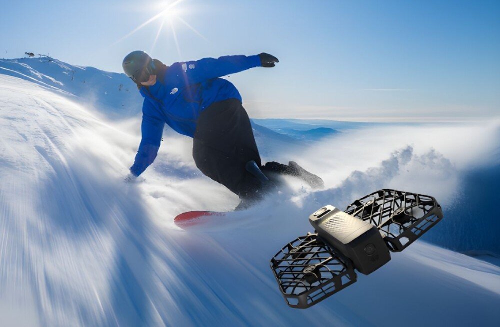 Dron HOVERAIR X1 Pro Basic Combo Snowboardzista w niebieskiej kurtce jadący po nasłonecznionym stoku z lecącym przed nim czarnym dronem, opis
