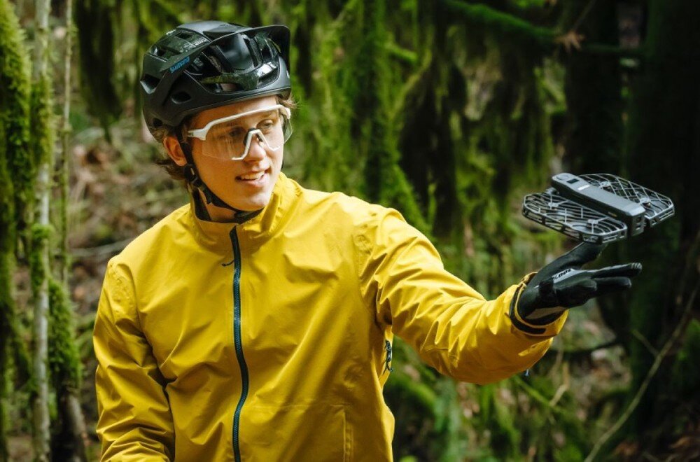 Dron HOVERAIR X1 Pro Ultimate Cycling Kit Snowboardzista w niebieskiej kurtce jadący po nasłonecznionym stoku z lecącym przed nim czarnym dronem, opis