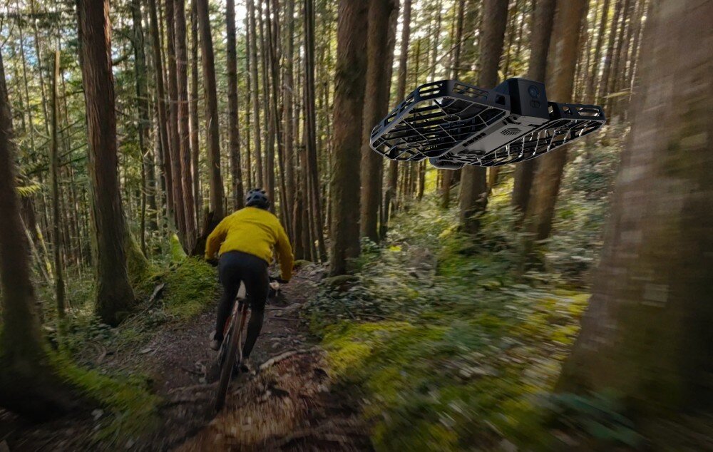 Dron HOVERAIR X1 Pro Ultimate Cycling Kit Rowerzysta jadący leśną ścieżką i lecący nad nim czarny dron, tryby, funkcje, ujęcia