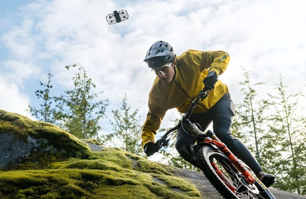 Dron HOVERAIR X1 Promax Snowboardzista w niebieskiej kurtce zjeżdżający po ośnieżonym stoku w pełnym słońcu, śledzony z boku przez niewielki dron w ochronnej kratownicowej obudowie, opis