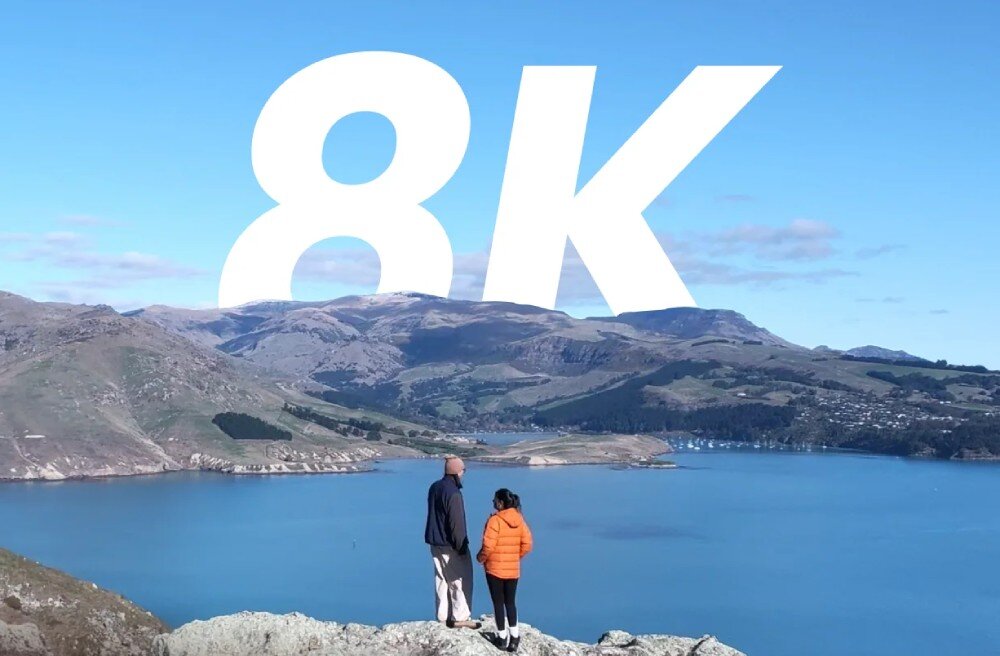 Dron HOVERAIR X1 Promax Panorama gór i jeziora w słoneczny dzień z dwiema osobami stojącymi na skale na pierwszym planie, nad krajobrazem dominuje duży biały napis 8K, nagrywanie, rozdzielczość, stabilizacja