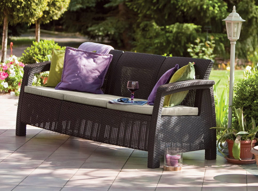 Sofa ogrodowa KETER Cofru Love Seat 3 osobowa Grafitowy sofa na tarasie obok latarnia w tle rośliny i drzewa z wysokiej jakości technorattanu odporny na uszkodzenia mechaniczne długa żywotność imitacja naturalnego splotu rattanowego sofa zachowa swój wygląd i funkcjonalność nie zblaknie nie popęka zabezpieczyć poduszki przed wilgocią i opadami
