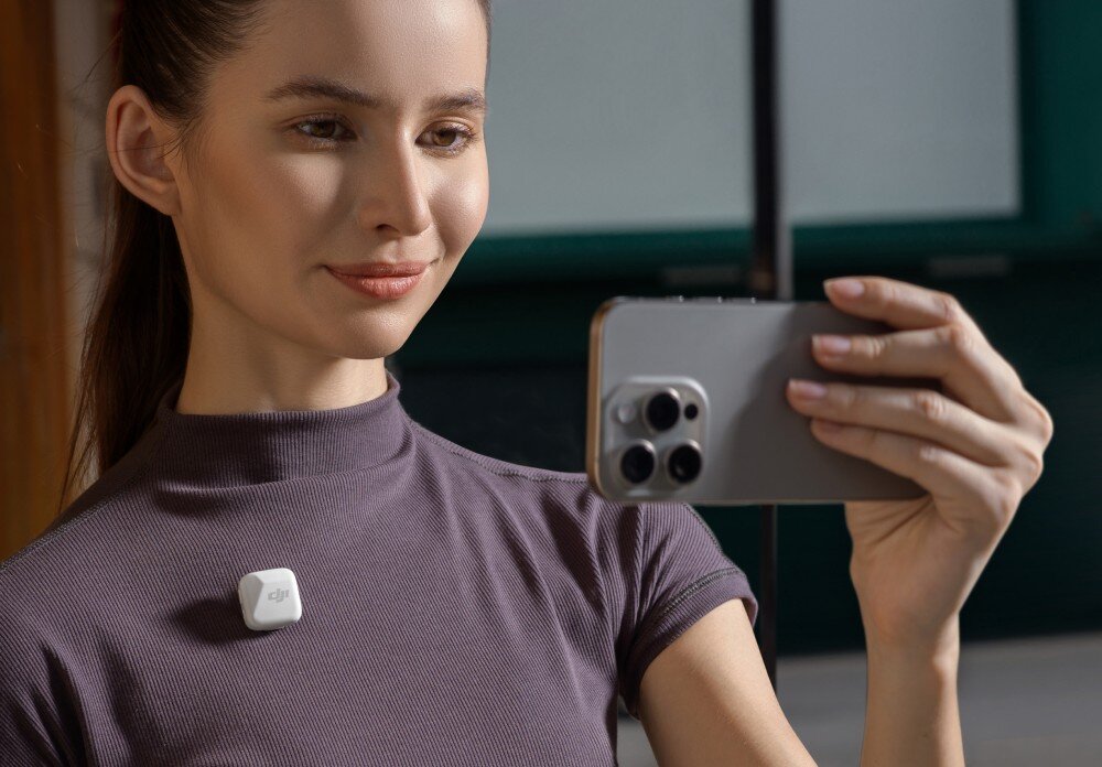 Osoba trzyma smartfon z trzema obiektywami aparatu, a na jej ubraniu przypięty jest biały mikrofon z logo DJI.
