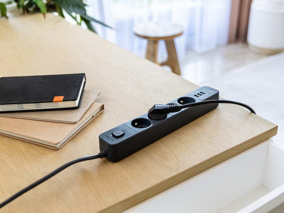 Na drewnianym blacie znajduje się czarna listwa zasilająca z kilkoma gniazdami i włącznikiem. Obok leżą dwa zeszyty, a w tle widać roślinę i stołek.