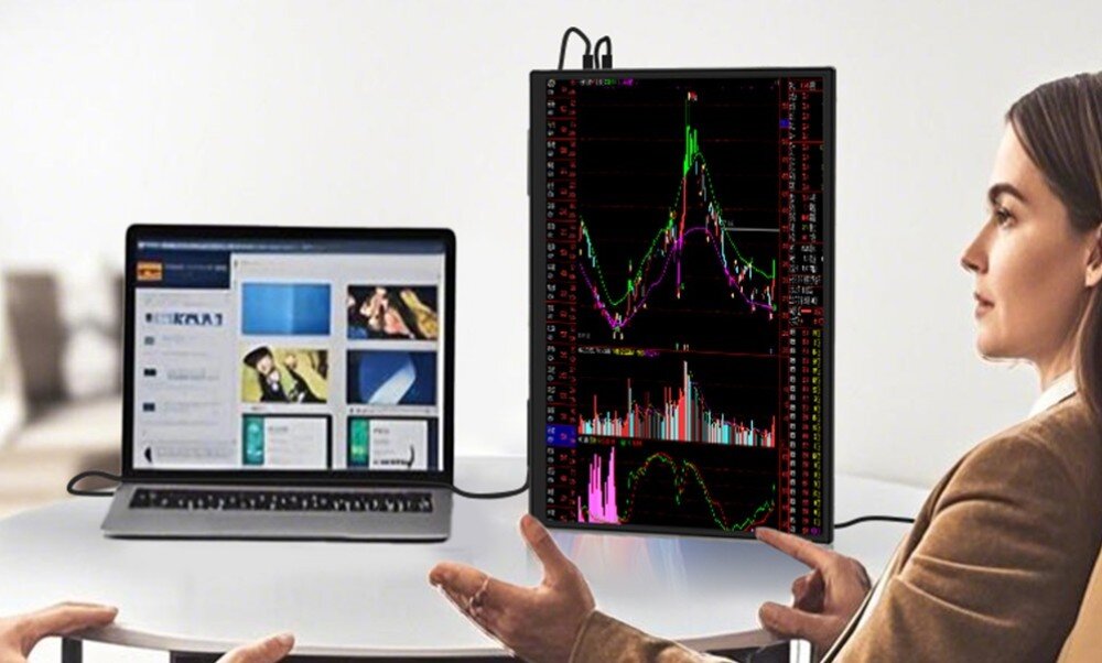 Monitor ZEUSLAP Z18S Kobieta siedząca przy stole obok otwartego laptopa, obok którego stoi monitor w orientacji pionowej wyświetlający kolorowe wykresy giełdowe, opis