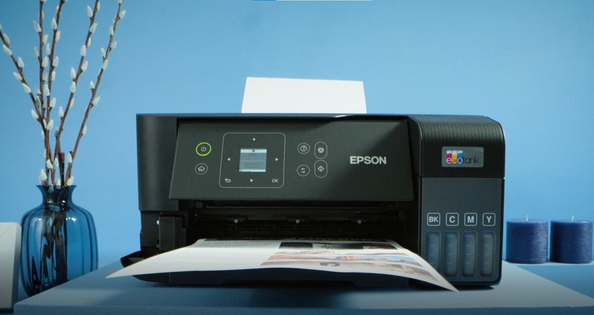 Na zdjęciu znajduje się czarne urządzenie wielofunkcyjne z logo EPSON i EcoTank, ustawione na niebieskim tle. Obok urządzenia widoczne są niebieskie świece i wazon z gałązkami.