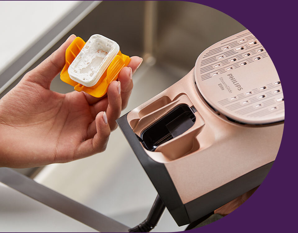 Osoba trzyma żółty pojemnik z białą substancją, obok znajduje się żelazko z widocznym napisem \'PHILIPS Azur 8000 Series\'.