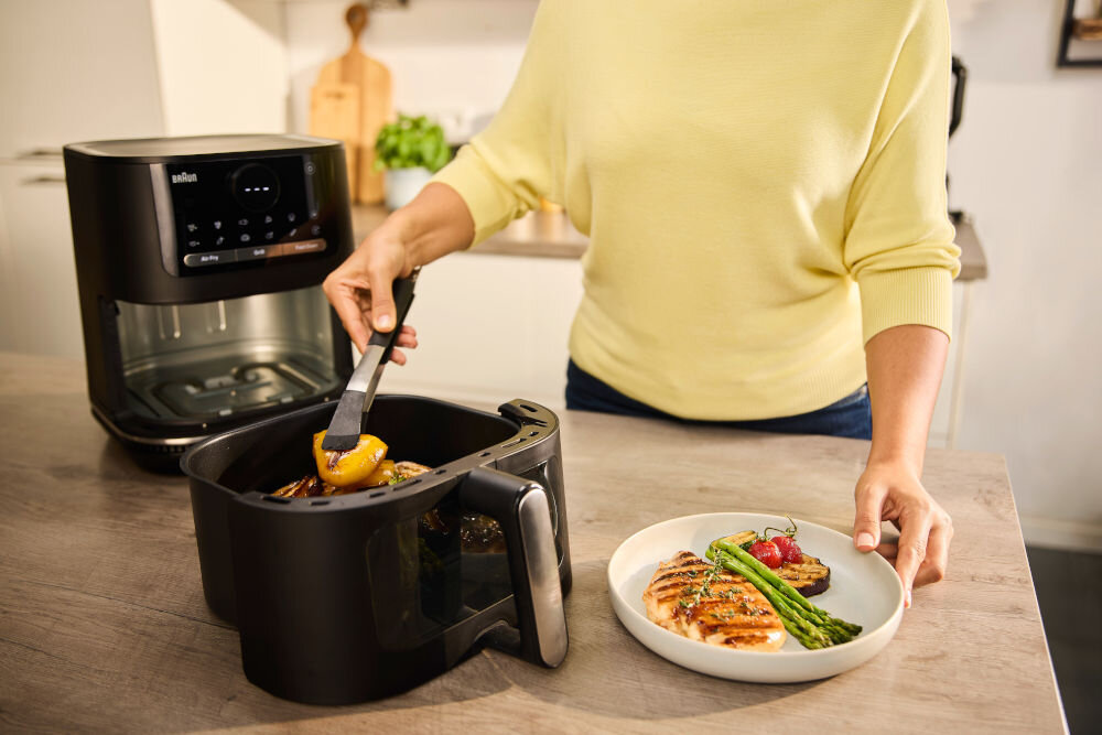Air Fryer Frytkownica beztłuszczowa BRAUN MultiFry 5 HF5073 frytkownica stoi na blacie osoba wyciąga z kosza potrawę na talerz z 20 automatycznymi programami gotowania zdrowe posiłki dla całej rodziny urządzenie małego AGD pełnowartościowe menu przygotowanie posiłków na zapas smażenie pieczenie grillowanie optymalne rezultaty podwójny element grzewczy nad i pod przetwarzanymi składnikami 