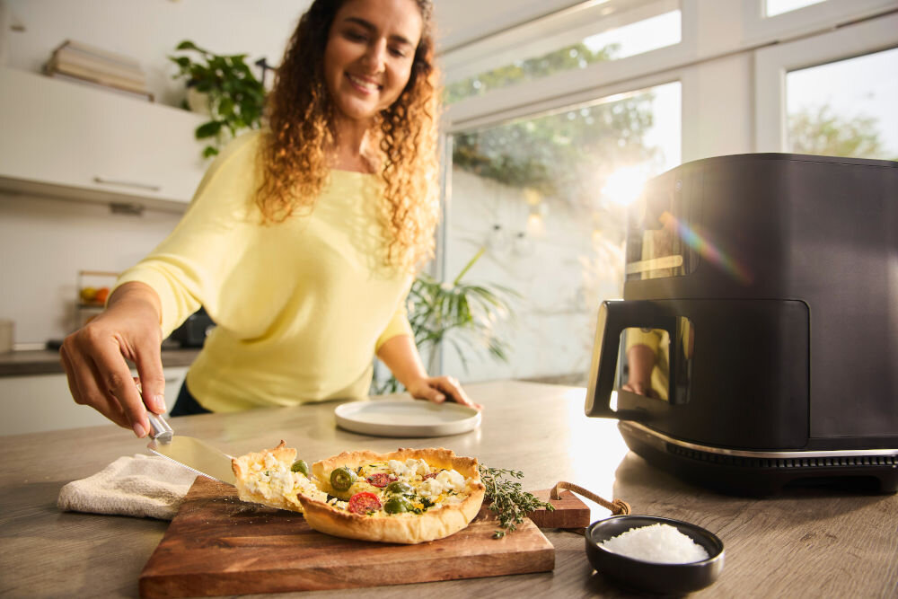 Air Fryer Frytkownica beztłuszczowa BRAUN MultiFry 5 HF5073 frytkownica stoi na blacie czarna obudowa panel przyciski uchwyt kobieta nakłada pokrojoną potrawę z deski z 20 automatycznymi programami gotowania oszczędność aż do 70% energii elektrycznej mniejsze rachunki za prąd świadomy wybór ochrona środowiska szybkie równomierne przygotowanie ulubionych potraw dla oszczędnych i ekologicznych gospodarstw domowych chrupiące frytki inne dania 