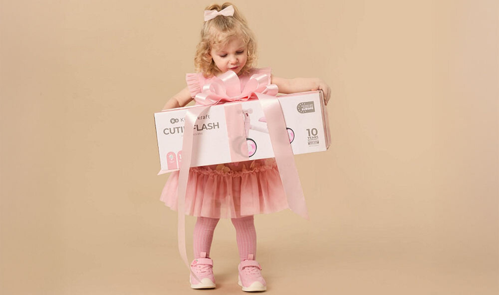 ROWEREK BIEGOWY KINDERKRAFT CUTIE FLASH BLUE dziewczynka trzyma zapakowane pudełko pomysł na prezent wsparcie rozwoju dziecka doskonalenie równowagi koordynacji ruchowej ogólna sprawność fizyczna podarunek na urodziny święta Dzień Dziecka mnóstwo frajdy stymulacja zdrowego rozwoju niezapomniane chwile pełne uśmiechu