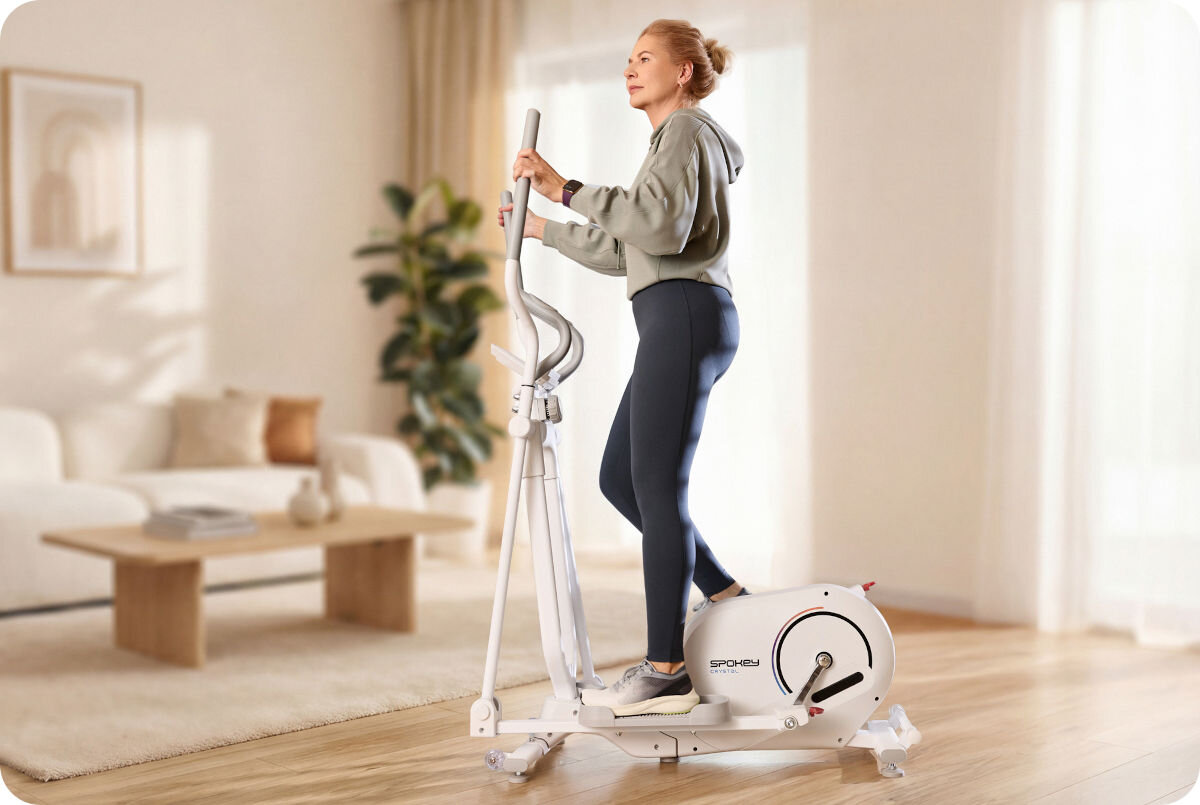 Orbitrek magnetyczny SPOKEY Crystal, kardio, aktywność, siła, wytrzymałość, kobieta na orbitreku ćwiczy w pokoju dziennym