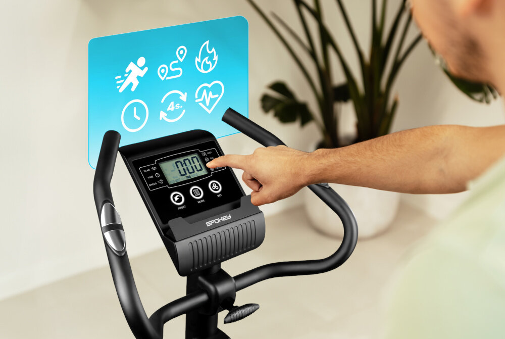Rower magnetyczny SPOKEY Dust pokój zbliżenie na wyświetlacz osoba siedzi na rowerze dotyka palcem wyświetlacza wbudowane sensory w kierownicy umożliwiają pomiar pulsu kontrola organizmu wyświetlacz LCD z funkcją SCAN śledzenie postępów motywuje do osiągania coraz lepszych rezultatów