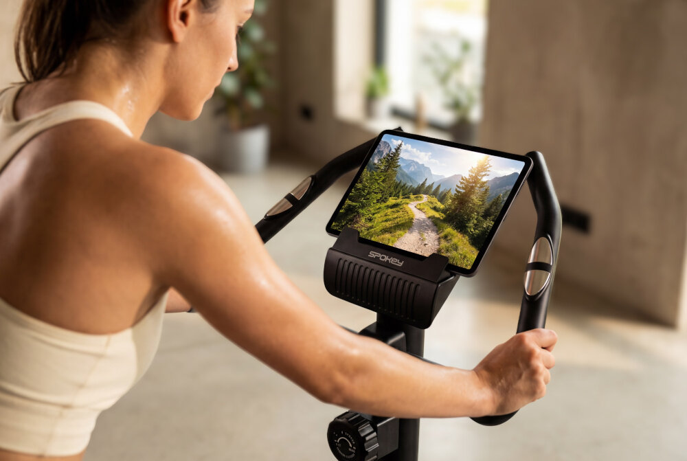 Rower magnetyczny SPOKEY Dust pokój osoba jedzie na rowerze ma tablet na uchwycie w tle ściana okno praktyczne miejsce na urządzenie mobilne smartfon lub tablet zawsze w zasięgu wzroku wygodne korzystanie z aplikacji treningowych kontrola parametrów jazdy zwiększa zaangażowanie podczas ćwiczeń trening jest przyjemniejszy bardziej motywujący