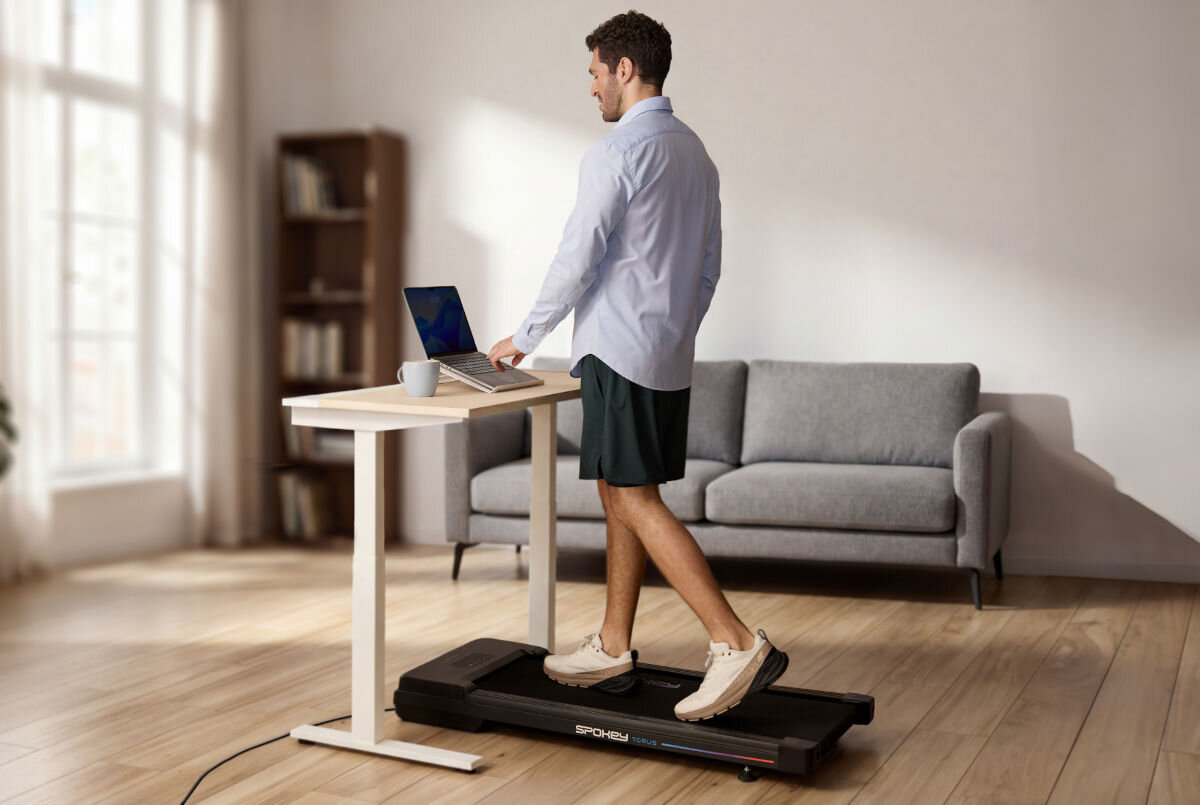 Bieżnia elektryczna SPOKEY Torus 946145 745.7W 6 km/h, ergonomia, mobilność, oszczędność, praktyczność, Mężczyzna w biurze chodzi na bieżni Spokey WALK pod biurkiem z laptopem.