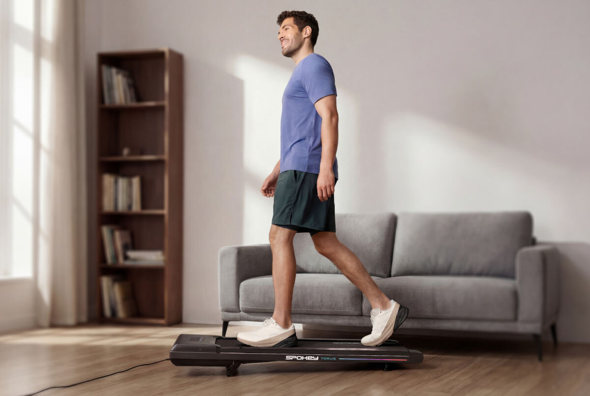 Bieżnia elektryczna SPOKEY Torus 946145 745.7W 6 km/h, utrzymanie, sprawność, serwis, trwałość, Mężczyzna w koszulce maszeruje na bieżni Spokey WALK obok regału z książkami.