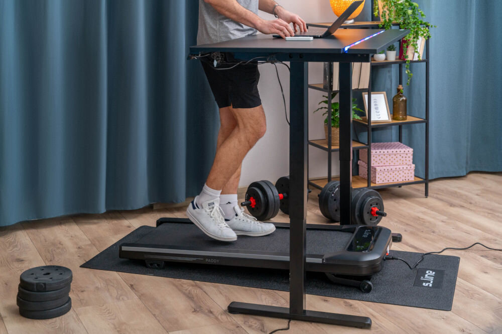 Bieżnia elektryczna S_LINE Paddy 920W 6 km/h Mężczyzna pracujący na laptopie przy biurku regulowanym podczas chodzenia na bieżni podbiurkowej S-line Paddy.
