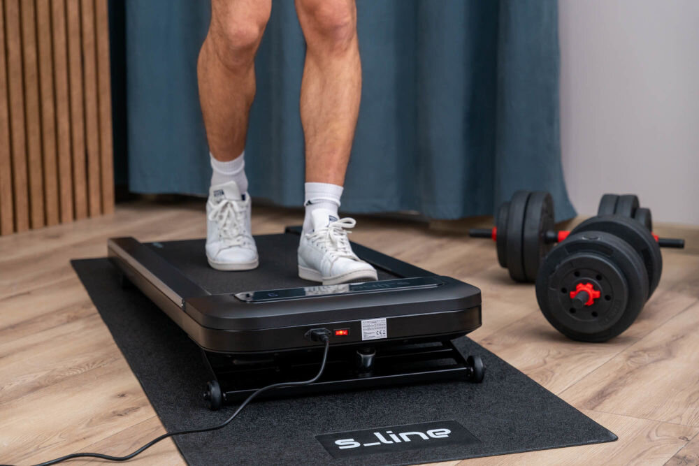 Bieżnia elektryczna S_LINE Paddy 920W 6 km/h Nogi mężczyzny podczas marszu na kompaktowej bieżni elektrycznej ustawionej na macie ochronnej w domowej siłowni.