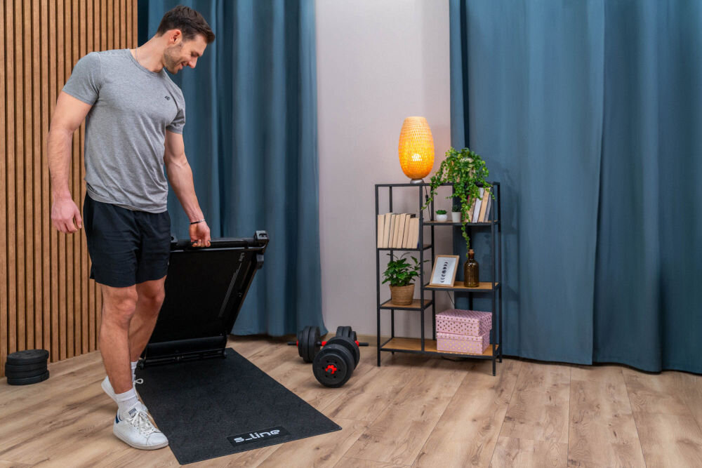 Bieżnia elektryczna S_LINE Paddy 920W 6 km/h Mężczyzna podnoszący lekką bieżnię podbiurkową S-line Paddy, prezentujący jej mobilność i łatwość przechowywania.
