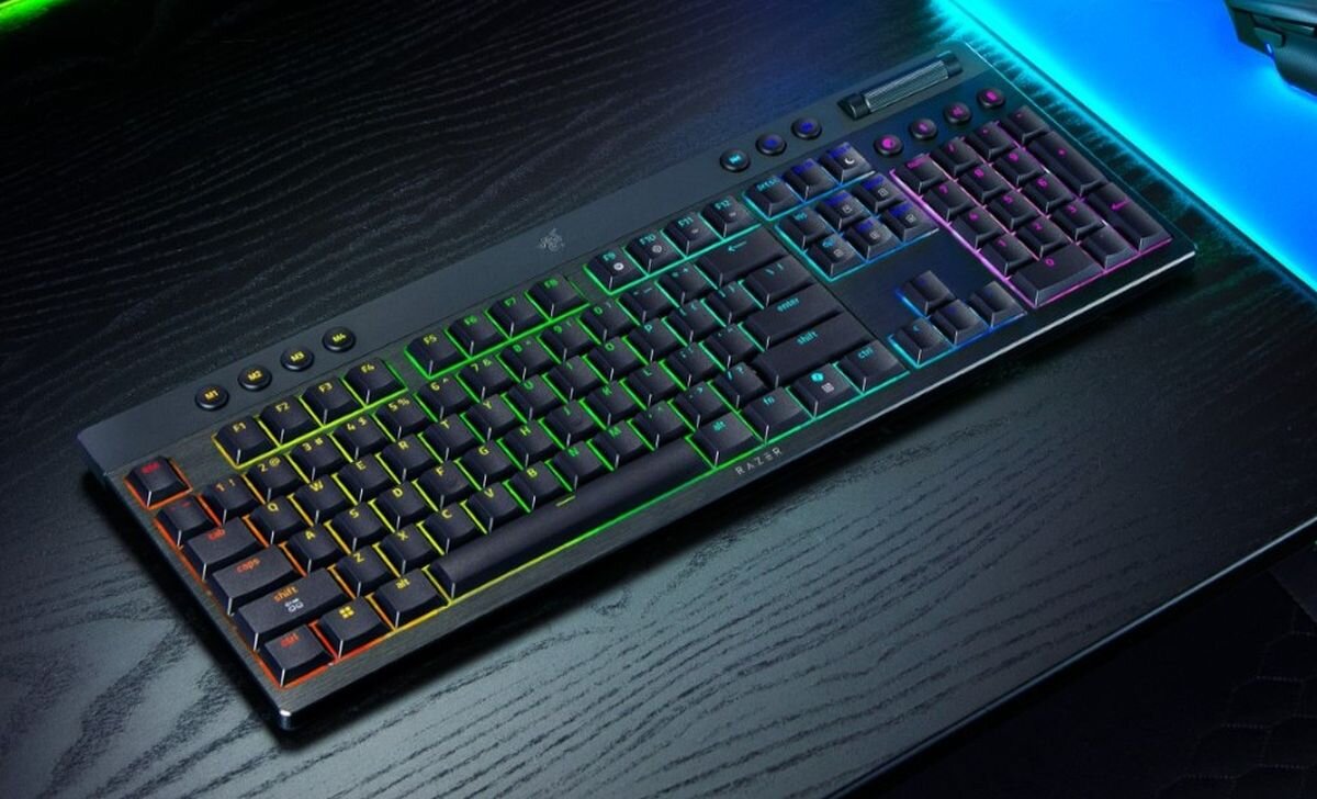 Na ciemnym biurku leży klawiatura z podświetleniem RGB, z widocznym logo producenta na środku. Klawisze są podświetlone w różnych kolorach, tworząc efekt tęczy.