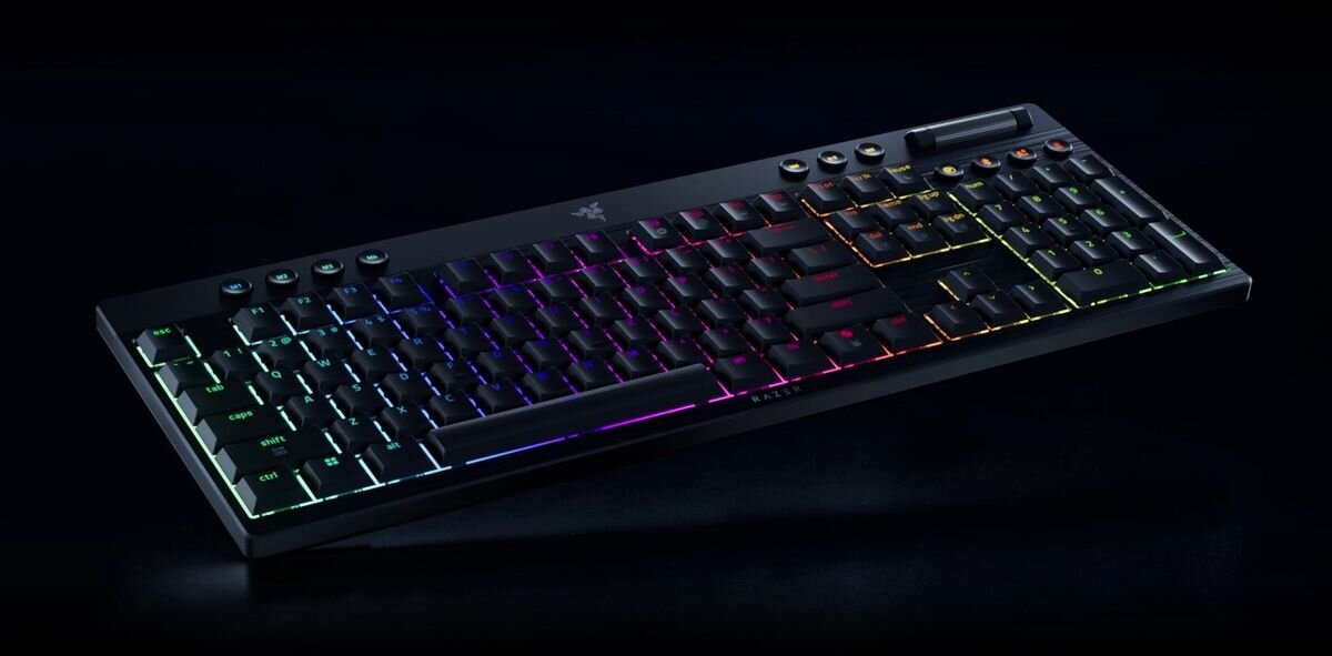 Klawiatura mechaniczna z podświetleniem RGB, widoczne logo producenta na środku. Klawisze mają różnokolorowe podświetlenie, a na górze znajdują się dodatkowe przyciski funkcyjne.