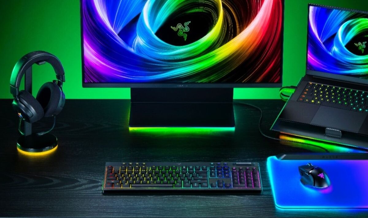 Na biurku znajduje się klawiatura z podświetleniem RGB, mysz na podkładce, słuchawki na stojaku oraz monitor i laptop z kolorowymi ekranami. Na monitorze widoczne jest logo Razer.