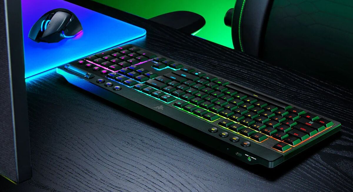 Na ciemnym biurku znajduje się klawiatura z podświetleniem RGB oraz myszka na podkładce. Na klawiaturze widoczne jest logo Razer.
