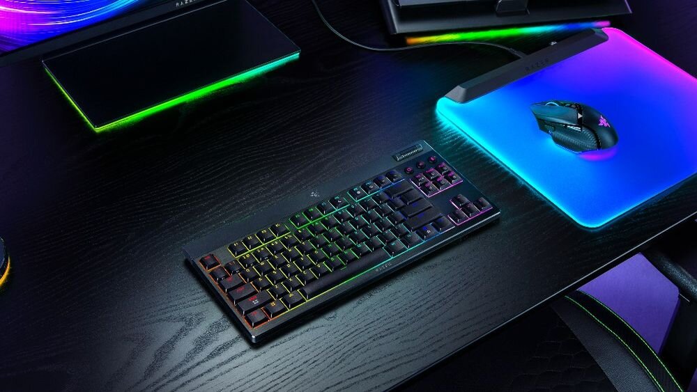 Na ciemnym biurku znajduje się klawiatura z podświetleniem RGB oraz mysz na podkładce z kolorowym podświetleniem. W tle widoczne są inne urządzenia elektroniczne z zielonymi akcentami świetlnymi.