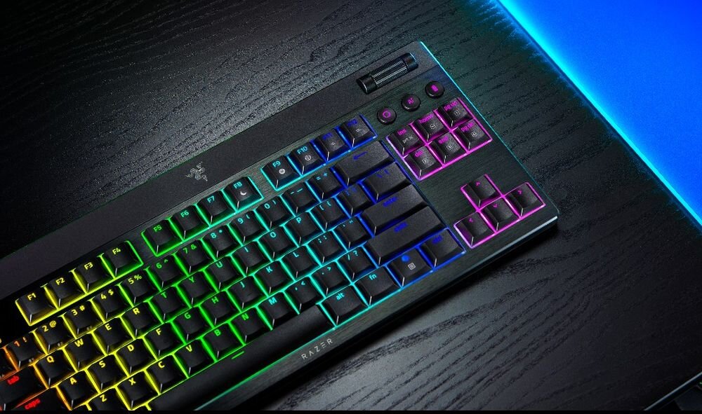 Na ciemnym biurku leży klawiatura z podświetleniem RGB, z widocznym logo Razer. Klawisze mają różne kolory, a na obudowie znajduje się napis \'RAZER\'.