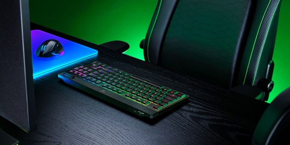 Na biurku znajduje się klawiatura z podświetleniem RGB oraz mysz na podkładce z efektem świetlnym. W tle widoczne jest krzesło gamingowe.