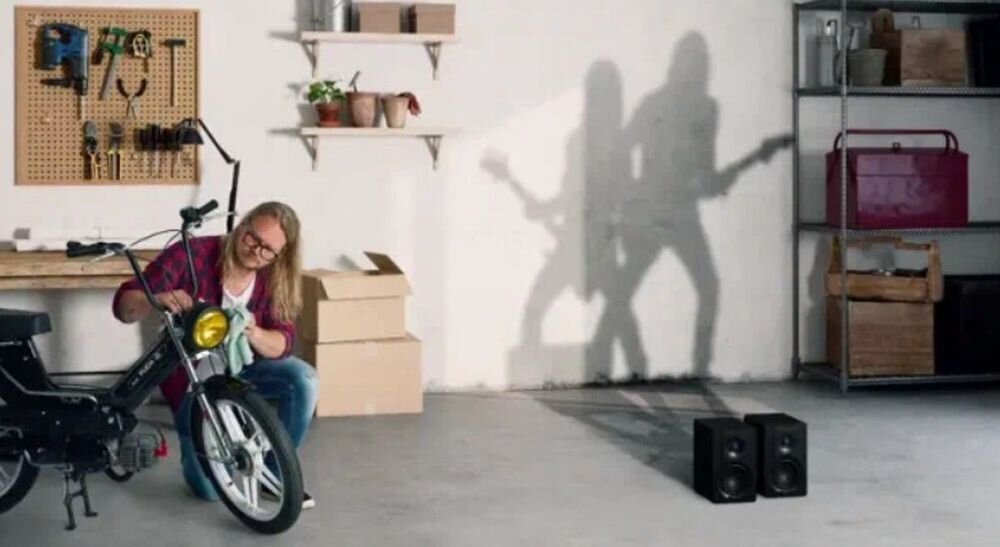 Kolumny głośnikowe DALI KUPID kompaktowe wymiary 24.5x15x19.8 cm ustawienie półka biurko, kolumny stojące na ziemi, osoba polerująca motocykl