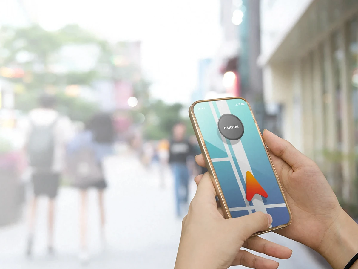 Osoba trzyma smartfon z aplikacją mapową, na ekranie widoczny jest lokalizator z napisem \'CANYON\'. W tle rozmyty widok ulicy z przechodniami.