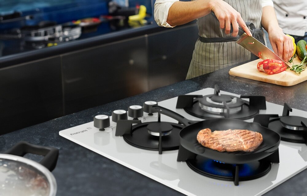 PŁYTA GAZOWA ISEASY MGBG-604B Osoba smaży stek na patelni ustawionej na jednym z palników, obok krojąc świeże warzywa na desce. Kuchenny blat w tle wypełniają akcesoria potrzebne do przygotowania posiłku palniki 3,3kW 1,75kW 1kW cztery strefy grzewcze regulacja mocy