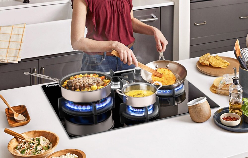 Płyta gazowa ISEASY MGBG-654A Na płycie gazowej jednocześnie smażone są różne potrawy, a garnki i patelnie stoją na trzech palnikach z mocnym, niebieskim płomieniem. Wokół ustawione są składniki i naczynia potrzebne do gotowania szklana płyta gazowa nowoczesny design trwałe materiały łatwa pielęgnacja