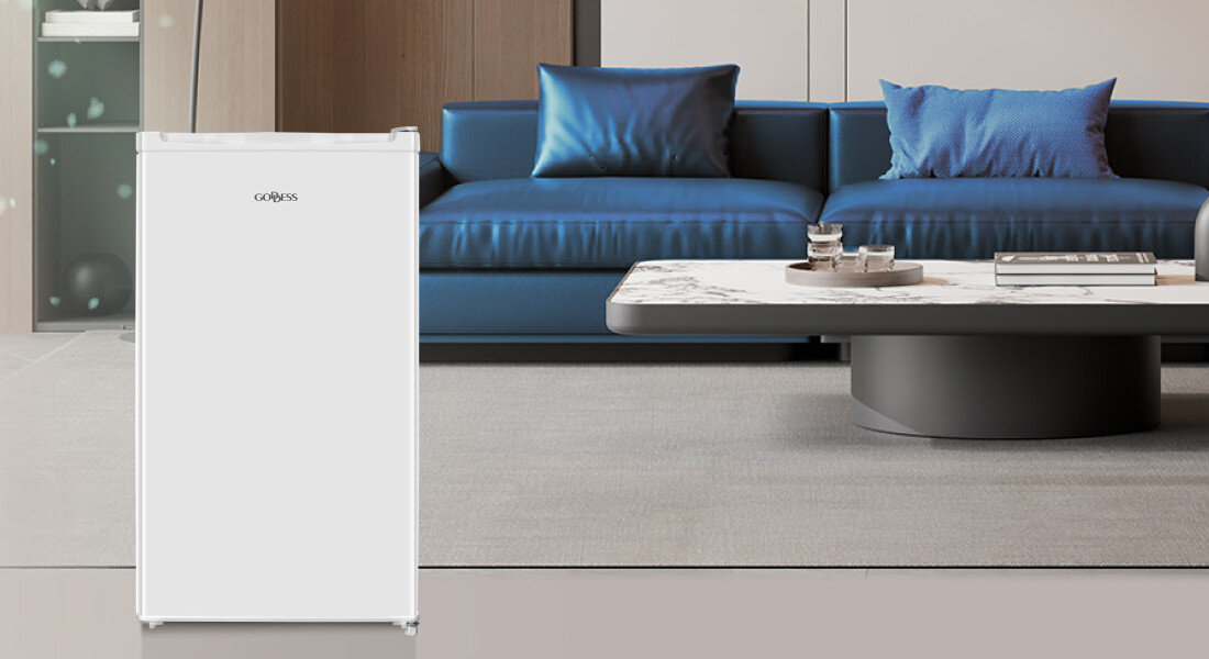 Lodówka GODDESS RME085GW8SSE Zdjęcie przedstawia białą lodówkę GOBESS ustawioną w salonie z niebieską sofą i niskim stolikiem kawowym. Minimalistyczny design urządzenia dobrze komponuje się z eleganckim wnętrzem