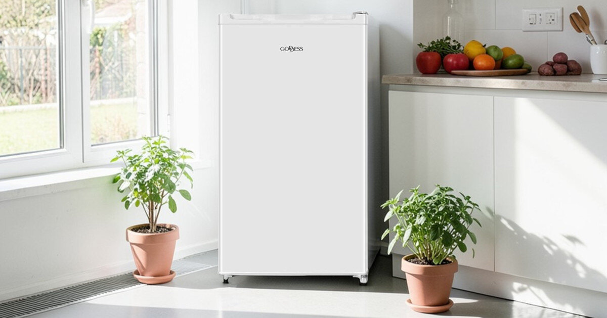 Lodówka GODDESS RME085GW8SSE Zdjęcie ukazuje białą lodówkę GOBESS stojącą w jasnej kuchni, obok blatu z owocami i doniczek z roślinami. Naturalne światło z okna podkreśla kompaktowe wymiary i prostą formę urządzenia lodówka kompaktowa mała przestrzeń kuchnia biuro kawalerka wymiary 85 × 48 × 45 cm