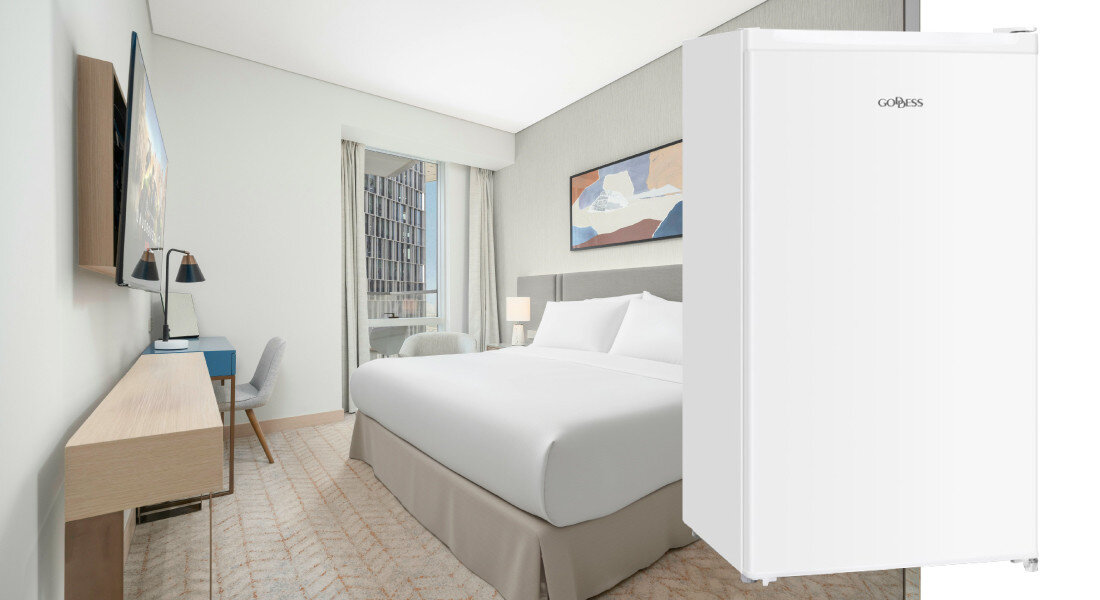 Lodówka GODDESS RME085GW8SSE Zdjęcie ukazuje białą lodówkę GOBESS w aranżacji sypialnianej lub hotelowej, obok łóżka i biurka. Kompaktowy rozmiar urządzenia podkreśla jego uniwersalne zastosowanie biała lodówka uniwersalny design lekka konstrukcja łatwy transport
