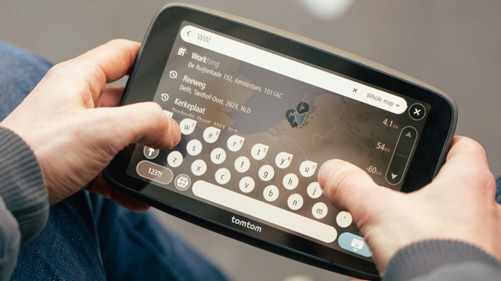 Osoba trzyma urządzenie nawigacyjne z widocznym ekranem dotykowym, na którym wyświetlana jest klawiatura i lista lokalizacji. Na dole ekranu widoczne jest logo \'tomtom\'.